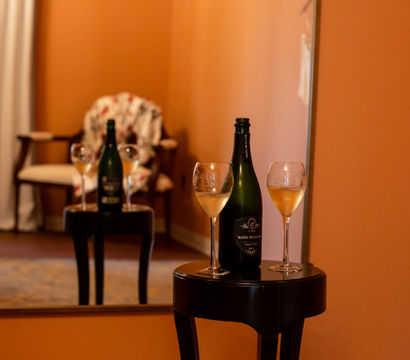 Bottle of sparkling wine and two filled wine glasses on a small black side table in a cozy hotel suite, reflected in a large mirror with a floral armchair and warm orange walls.