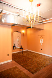 Peach-colored dressing room interior with a gold chandelier, exposed HVAC ductwork, large ornate mirror, small table with wine bottle and glass, white lingerie hung on the wall, and brown patterned rugs over hardwood floor.
