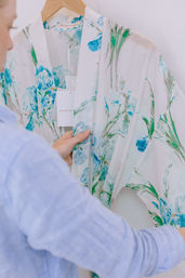 Close-up of a person holding a white robe with blue-green watercolor floral print on a wooden hanger, tag dangling over soft, breezy fabric.