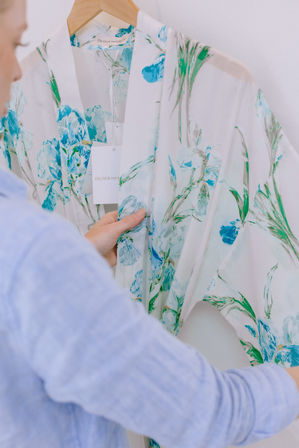 Close-up of a person holding a white robe with blue-green watercolor floral print on a wooden hanger, tag dangling over soft, breezy fabric.