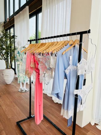 Sunlit boutique display: pastel silk robes and lace lingerie (blue, pink, white) on wooden hangers along a black clothing rack with hardwood floors and large windows.