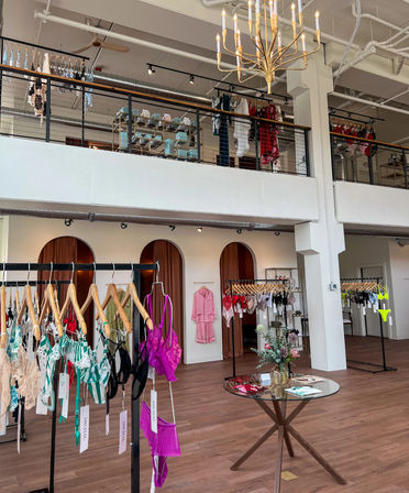 Loft-style lingerie boutique showroom with two-story industrial-chic interior, chandelier, wooden floors, arched fitting-room curtains, glass display table with flowers, and colorful bras and underwear on racks and mezzanine.