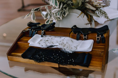 Elegant beaded white and black bridal garters and black suspenders arranged on a wooden tray on a glass table — wedding accessories styling shot with a soft floral arrangement in the background.