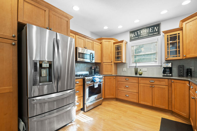 Sunny modern kitchen with stainless steel French-door refrigerator and gas range, warm maple cabinets, granite countertops, hardwood floor, coffee maker, and decorative "KITCHEN" sign above the window.