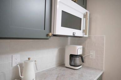 Modern kitchen countertop with white over-the-range microwave, white drip coffee maker and gooseneck kettle, gray shaker cabinet with brass hardware, and pale square tile backsplash