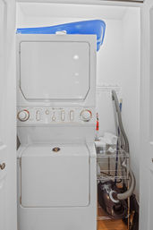 Compact interior laundry closet with stacked white washer-dryer, blue plastic laundry basket on top, metal wire shelving with cleaning supplies and a canister vacuum