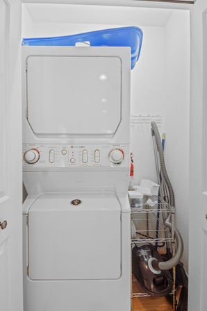 Compact interior laundry closet with stacked white washer-dryer, blue plastic laundry basket on top, metal wire shelving with cleaning supplies and a canister vacuum