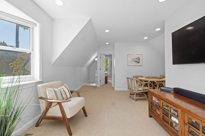 Bright attic loft living room with sloped ceilings and recessed lighting, beige armchair by a sunny window, round rattan dining table with chairs, wall-mounted TV and wooden media console.