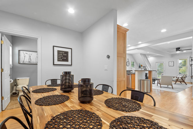 Open-plan dining area and kitchen in a modern suburban home with a wooden farmhouse table, black woven placemats and lanterns, metal chairs, light oak floors and bright living room beyond.
