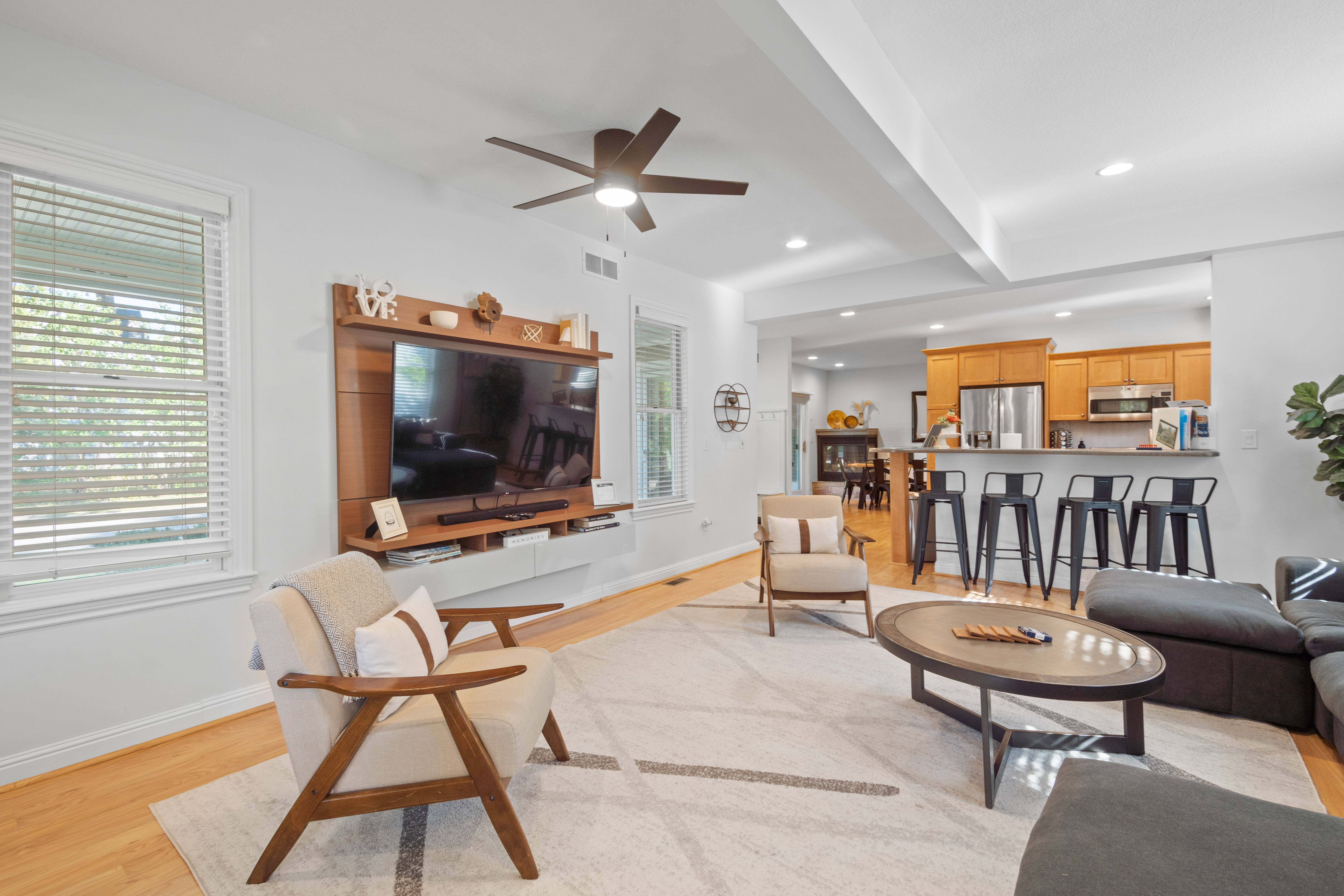 Cozy bright open-concept living room and kitchen with hardwood floors, modern ceiling fan, wall-mounted TV on a wooden media wall, mid-century armchairs, round coffee table, and a breakfast bar with black metal stools.