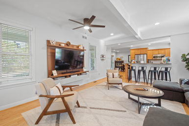 Cozy bright open-concept living room and kitchen with hardwood floors, modern ceiling fan, wall-mounted TV on a wooden media wall, mid-century armchairs, round coffee table, and a breakfast bar with black metal stools.
