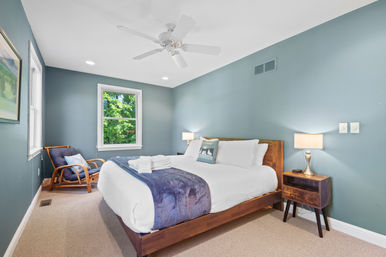 Cozy bedroom interior with teal walls, wooden platform bed dressed in white linens and a navy throw, bedside lamp on a wooden nightstand, rattan armchair, ceiling fan, and a window showing green trees.