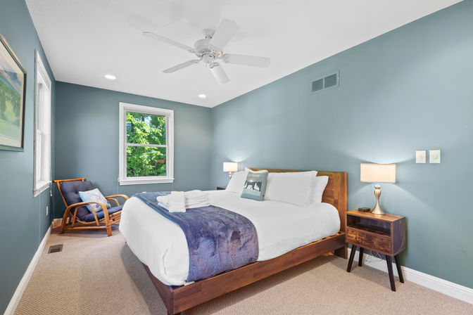 Cozy bedroom interior with teal walls, wooden platform bed dressed in white linens and a navy throw, bedside lamp on a wooden nightstand, rattan armchair, ceiling fan, and a window showing green trees.