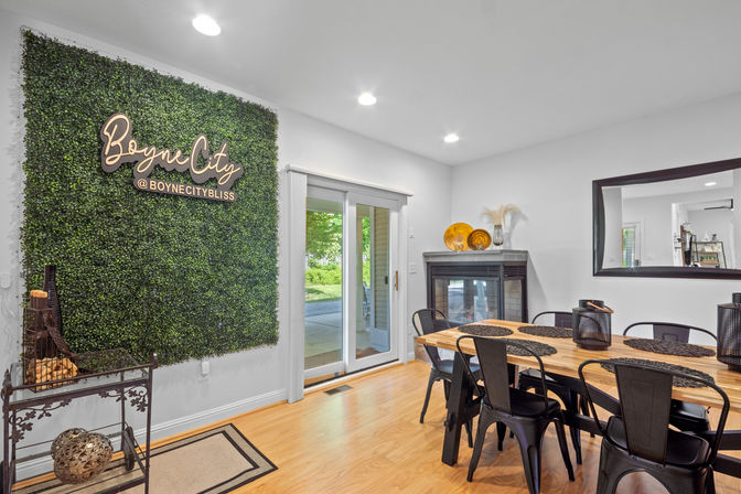 Cozy modern dining room with a large faux boxwood accent wall and cursive sign, sliding glass patio doors, wooden dining table surrounded by black metal chairs with lantern centerpieces, a fireplace topped with decorative plates, and a large wall mirror.