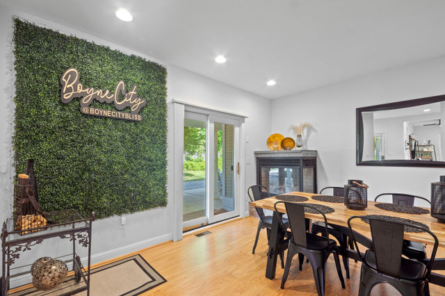 Cozy modern dining room with a large faux boxwood accent wall and cursive sign, sliding glass patio doors, wooden dining table surrounded by black metal chairs with lantern centerpieces, a fireplace topped with decorative plates, and a large wall mirror.