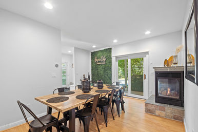 Bright modern dining room with a long wooden farmhouse table, black metal chairs and woven placemats, indoor gas fireplace with stone hearth, sliding glass doors to the yard and a green living wall with neon accent.