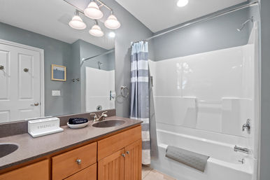 Bright modern gray bathroom interior with wood vanity and dark countertop, sink and chrome faucet, large mirror with overhead lights, and a white tub‑shower with a gray-and-white striped curtain