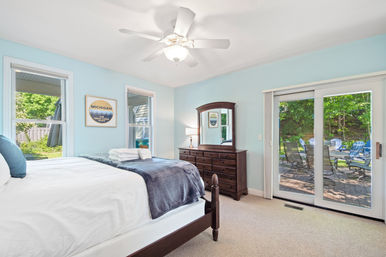 Bright lakeside-style bedroom with pale blue walls, white ceiling fan, wooden dresser and mirror, neatly made bed with folded towels, and sliding glass doors opening to a patio with blue Adirondack chairs