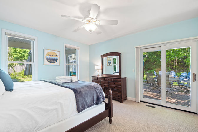 Bright lakeside-style bedroom with pale blue walls, white ceiling fan, wooden dresser and mirror, neatly made bed with folded towels, and sliding glass doors opening to a patio with blue Adirondack chairs
