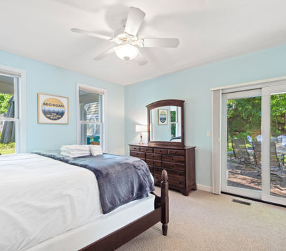 Bright lakeside-style bedroom with pale blue walls, white ceiling fan, wooden dresser and mirror, neatly made bed with folded towels, and sliding glass doors opening to a patio with blue Adirondack chairs