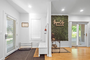 Welcoming entryway with a white bench and shoe rack, coir doormat, faux boxwood accent wall topped by a decorative script sign, sliding glass door to a sunny backyard, and warm hardwood floors.