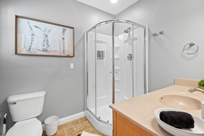 Modern gray bathroom with curved glass corner shower, beige vanity countertop, white toilet, and framed botanical print.