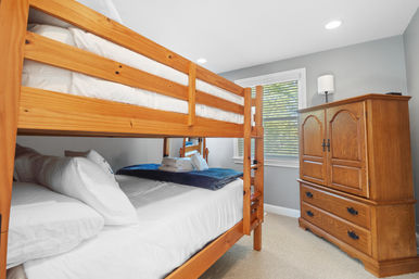 Cozy pine bunk bed in a bright guest bedroom with crisp white linens, gray walls, window blinds and a wooden armoire — perfect family stay setup.