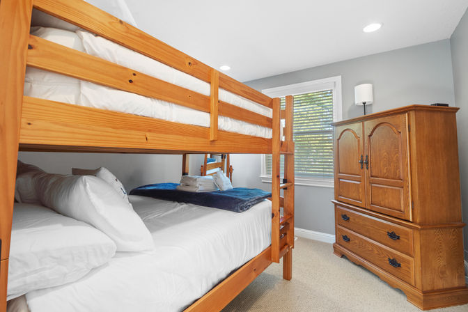 Cozy pine bunk bed in a bright guest bedroom with crisp white linens, gray walls, window blinds and a wooden armoire — perfect family stay setup.