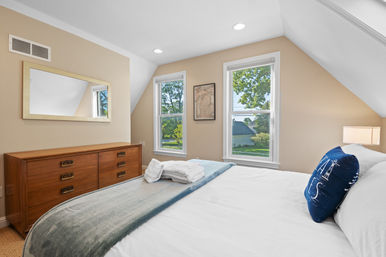 Cozy sunlit bedroom with beige walls and sloped ceiling, two tall windows overlooking a leafy suburban street, wooden dresser with mirror, neatly made bed with folded towels and a navy nautical pillow.