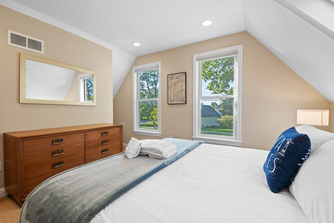 Cozy sunlit bedroom with beige walls and sloped ceiling, two tall windows overlooking a leafy suburban street, wooden dresser with mirror, neatly made bed with folded towels and a navy nautical pillow.
