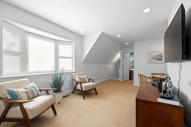 Sunlit attic sitting room with sloped ceiling and recessed lights, large bay window with light-filtering shades, two mid-century wooden armchairs with striped pillows, potted plant, carpeted floor, wall-mounted TV and wooden console topped with books and a small horse sculpture.