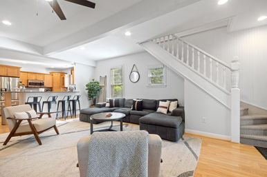 Bright open-concept living room and kitchen with a gray sectional sofa, round coffee table, white staircase, wood floors, and a breakfast bar with black metal stools.
