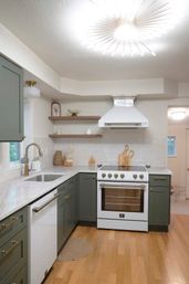 Cozy modern kitchen with sage-green lower cabinets, white range and hood, marble-look countertops, brass faucet, open wood shelves and warm hardwood floors.