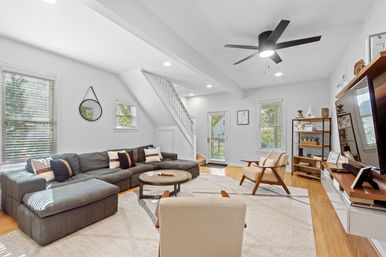 Cozy bright modern living room with gray sectional sofa, round coffee table, beige accent chairs, hardwood floors, black ceiling fan, windows with blinds and a white staircase.