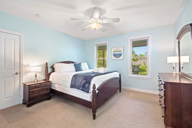 Cozy light-blue bedroom with dark wood bed and dresser, white linens and folded towels, ceiling fan overhead, two windows with backyard view and framed Michigan wall art.