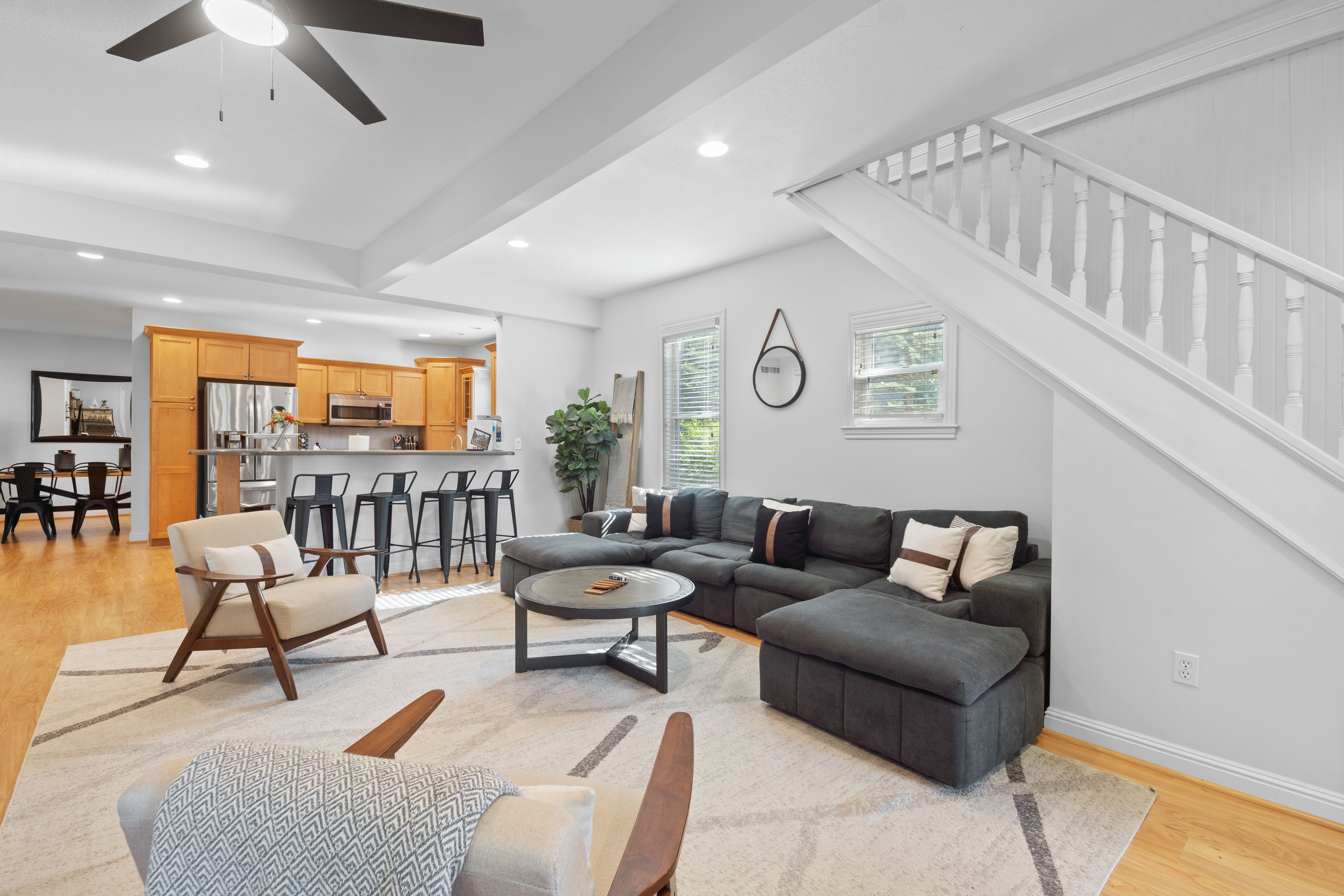 Bright open-concept living area and kitchen with gray sectional sofa, round coffee table, kitchen island with black bar stools, light hardwood floors, white staircase, ceiling fan and natural light.