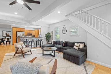 Bright open-concept living area and kitchen with gray sectional sofa, round coffee table, kitchen island with black bar stools, light hardwood floors, white staircase, ceiling fan and natural light.