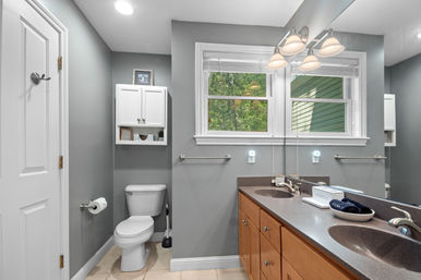 Bright modern gray bathroom in a home with double-sink vanity and wood cabinets, large mirror, toilet, white wall cabinet, towel bar, and a window showing leafy green trees.