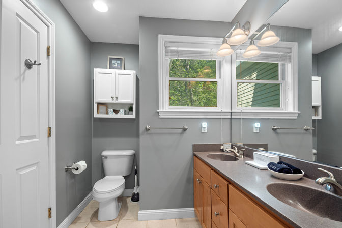 Bright modern gray bathroom in a home with double-sink vanity and wood cabinets, large mirror, toilet, white wall cabinet, towel bar, and a window showing leafy green trees.