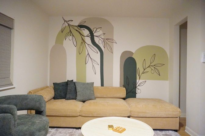 Cozy modern living room interior with a mustard velvet sectional, green velvet armchair, round coffee table and a painted abstract arch mural featuring leafy line art on the wall.