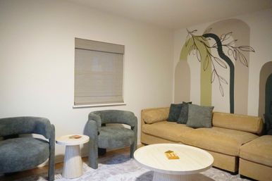Cozy modern living room with mustard-beige sofa, two rounded teal armchairs, light wood round coffee and side tables, neutral window blind, and an abstract arched wall mural with a leafy branch.