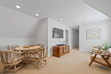 Cozy attic lounge with rattan dining set, beige carpet, wall-mounted TV, recessed ceiling lights and framed map art — beachy neutral decor.