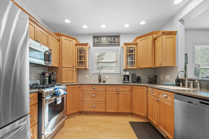 Sunny U-shaped kitchen with light maple cabinets, stainless-steel fridge and gas range, herringbone tile backsplash, recessed lighting, coffee station and a decorative "KITCHEN" sign above the window.