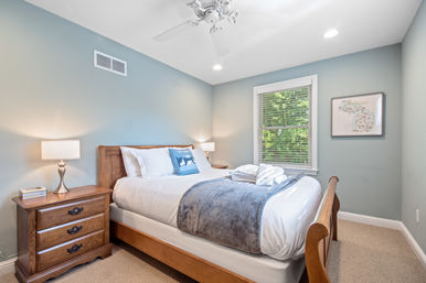 Cozy sunlit bedroom with wooden bed and nightstand, white linens and blue throw, folded towels, ceiling fan and bedside lamps against blue-gray walls with a window and framed Michigan map.