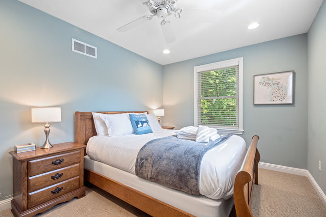 Cozy sunlit bedroom with wooden bed and nightstand, white linens and blue throw, folded towels, ceiling fan and bedside lamps against blue-gray walls with a window and framed Michigan map.