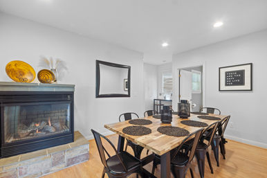 Cozy modern farmhouse dining room with a wooden table for six, black metal chairs, woven placemats and lantern centerpieces, a gas fireplace topped with amber decorative bowls, mirror and framed sign on light gray walls.