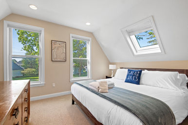 Sunny cozy attic bedroom with a skylight and two large windows, crisp white bedding, a navy "Great Lakes" pillow, folded towels, and leafy suburban views