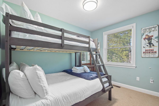 Cozy teal bedroom featuring a dark wood bunk bed with white linens and folded towels, ladder, window with blinds showing leafy outside view, ceiling light and vintage ski poster on the wall.