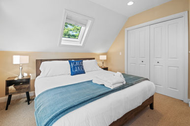 Cozy attic bedroom with angled skylight revealing green treetops, crisp white bedding, blue throw and lake-themed accent pillow, wooden nightstands with lamps, and white double closet doors.