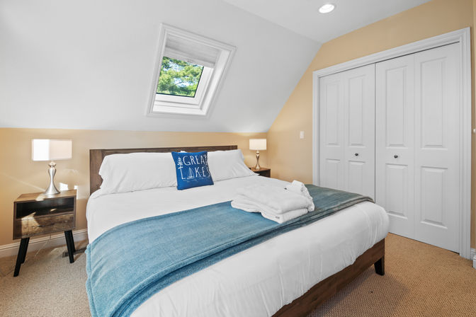 Cozy attic bedroom with angled skylight revealing green treetops, crisp white bedding, blue throw and lake-themed accent pillow, wooden nightstands with lamps, and white double closet doors.